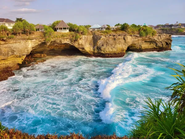 Stunning coastal view in Bali, Indonesia featuring turquoise waves crashing against rocky cliffs adorned with lush greenery and picturesque cottages.