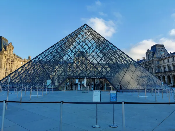 Louvre Museum glass pyramid entrance in Paris, France, showcasing iconic architecture and visitor queues against a blue sky.
